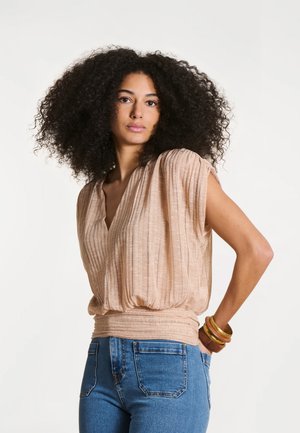 Femme aux cheveux bouclés volumineux portant un haut sans manches beige texturé et un jean bleu, posant avec une main sur la hanche et des bracelets.