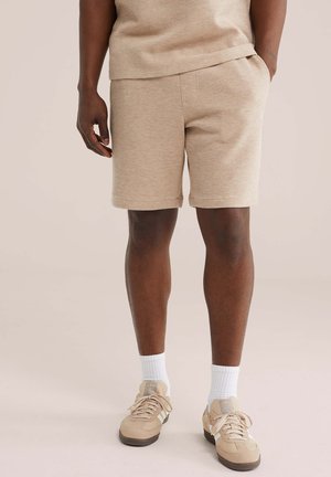 Moški v bež teksturiranih kratkih hlačah, usklajeni majici, belih nogavicah in bež Adidas supergah stoji na nevtralnem ozadju.