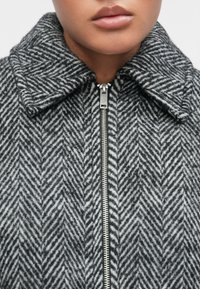 Veste en laine mélangée gris et noir à motif chevrons, dotée d'une fermeture éclair argentée et d'un large col, mettant en valeur le détail du tissu texturé.