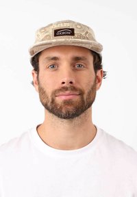 Casquette en tissu beige à motifs, avec une visière plate et un patch logo rectangulaire marron à l'avant. Portée par une personne en chemise blanche.