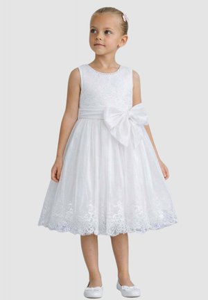 Ragazza giovane con vestito bianco senza maniche in pizzo con grande fiocco in vita e scarpe bianche abbinate, capelli legati con una molletta rosa.