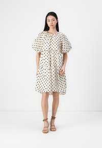 Krämdress med svarta prickar, puffärmar, flerlagat nederkant och avspänt snitt. Modellen bär sandaler med remmar i tan.