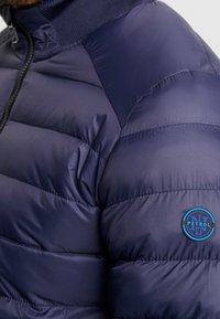 Námořnicky modrá prošívaná bunda s vysokým límcem a modrým kulatým logem na rameni, vyrobená z hladké, reflexní tkaniny.