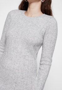 Donna che indossa un maglione a costine grigio chiaro con maniche lunghe e scollatura rotonda, mostrata dalla gola ai fianchi.