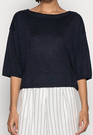 Kobieta ubrana w luźną ciemnogranatową bluzkę z rękawami do łokcia, zestawioną z białymi spodniami z wysokim stanem, ozdobionymi cienkimi pionowymi czarnymi paskami.