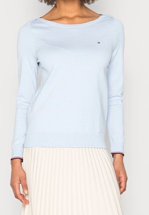 Kobieta ubrana w jasnoniebieski sweter z długim rękawem oraz białą plisowaną spódnicę, z małym logo na klatce piersiowej swetra i mankietach rękawów.