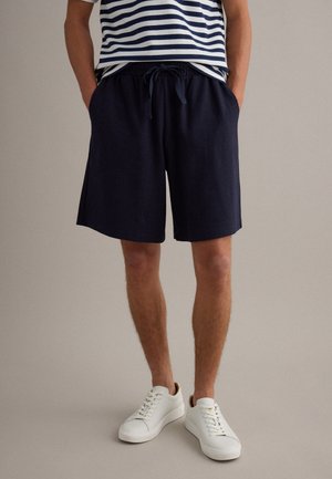 Man draagt marineblauwe short met trekkoord, witte sneakers en een marineblauw-wit gestreept shirt, staat met handen in de zakken.