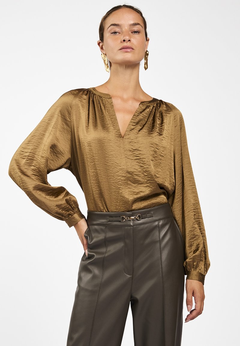 Bruine satijnen blouse met een V-hals, geraffineerd detail bij de schouders en lange mouwen. Gecombineerd met hooggewaaid groene neplederen broek.