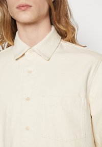 Camicia button-up color crema realizzata in tessuto testurizzato, con colletto a punta, taschino frontale e bottoni chiari.