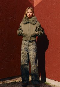 Olive zelena bomber jakna s umetnim krznom, kombinirana s svetlim denim hlačami z vzorcem leoparda. Postavljena pred teksturirano rdečo steno.