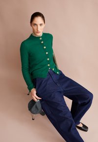 Vrouw met blauwe ogen die een groene button-up trui, een beige coltrui, losse donkerblauwe broek en zwarte schoenen draagt, zittend op een stoel en een grijze pet vasthoudend.