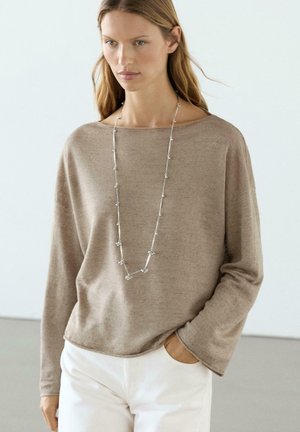 Donna che indossa un top beige a maniche lunghe e pantaloni bianchi, accessoriata con una lunga collana a catena d'argento, mano in tasca, sfondo neutro.