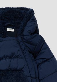 Marineblauwe pufferjack met een Sherpa-voering in de capuchon, voorzien van een gequiltde textuur en een label met de tekst "s.Oliver." Zacht materiaal voor warmte.