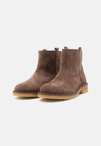 Botas de tornozelo de camurça castanha com puxadores, solas de crepe empilhadas baixas e costura visível sobre um fundo branco.