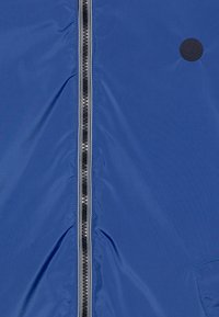 Giacca leggera blu con una cerniera nera verticale, tessuto testurizzato e un'etichetta rotonda con logo sul lato sinistro.