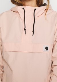 Jasnoróżowa kurtka z kapturem ze czarnymi sznurkami, kieszenią z przodu oraz szaro-czarną łatą z logo. Gładka faktura.