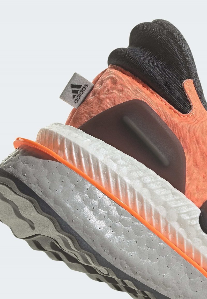 Parte superior de tela naranja con acentos negros, suela texturizada gris y franja media naranja; cuenta con una etiqueta del logo de Adidas en el talón.