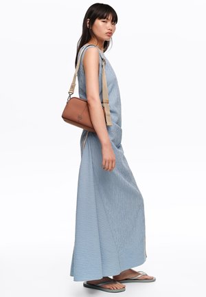 Mujer con un vestido maxi azul sin mangas, llevando un bolso marrón cruzado con correa beige, con chanclas, de pie frente a un fondo blanco.