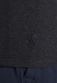 Zblízka černá žebrovaná látka s vyšívaným logem Polo Ralph Lauren nad modrým texturovaným pasem.