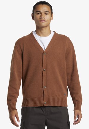 Cardigan marrón de punto con escote en V, que cuenta con un cierre de botones en la parte delantera y puños y dobladillo acanalados. Usado sobre una camiseta blanca.