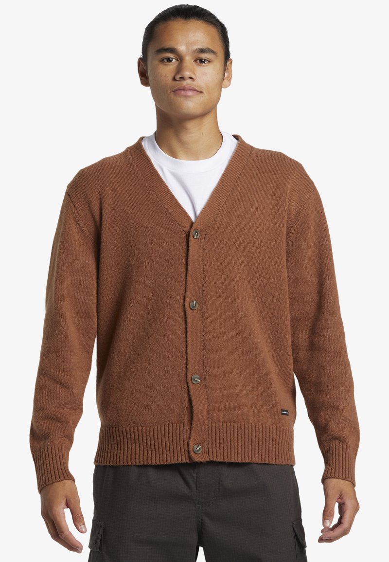 Brauner Strickcardigan mit V-Ausschnitt, mit Knopfverschluss vorne und gerippten Bündchen sowie Saum. Getragen über einem weißen T-Shirt.