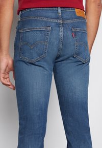 Mørkeblå denimjeans med rette ben, fem lommer og distinkte gule sømmer. Har et lærpatch på baksiden av livet.