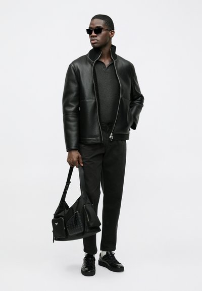 Črna usnjena jakna z zadrgo spredaj, siva polo majica, črne hlače in črni supergi. Drži črno torbico z okraski.