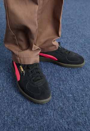 Osoba nosząca czarne i czerwone buty sportowe Puma Palermo z brązowymi spodniami stoi na niebieskim wykładzinowym podłodze.