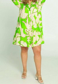 Groen en crèmekleurige jurk met bloemenprint, knopen aan de voorkant, gelaagd design, korte mouwen en een wijd uitlopende onderkant. Gecombineerd met lichtgekleurde sandalen.