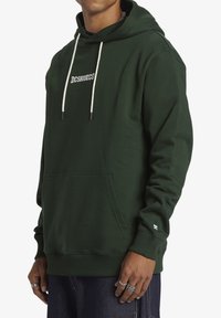 Felpa verde in misto cotone, con tasca a marsupio frontale, cordini con punte nere e logo "DCSHOECO" sul petto.