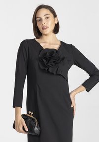 Czarna sukienka z długimi rękawami i głębokim dekoltem w szpic, z dużym falbaniastym kwiatowym akcentem na piersi. Trzyma czarną kopertówkę ze złotymi akcentami.