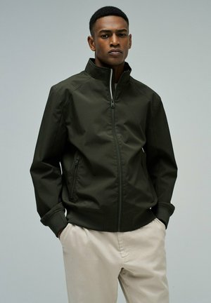 Hombre con chaqueta verde oscuro con cremallera y bolsillos con cremallera, y pantalones beige, de pie con las manos en los bolsillos contra un fondo liso.