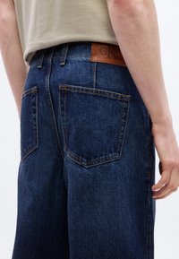 Άτομο που φοράει σκούρο μπλε τζιν με πίσω τσέπες και καφέ δερμάτινο λογότυπο Calvin Klein στη μέση.