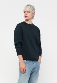 Hanorac bleu marin cu guler rotund, cu manșete și tiv din material elastic, având un logo discret pe piept. Purtați-l cu blugi deschiși la culoare.
