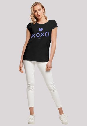 Schwarzes T-Shirt mit kurzen Ärmeln, das den Text "I ❤️ XOXO" in helllila zeigt. Kombiniert mit weißen, figurschmeichelnden Hosen und weißen Sneakers.