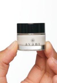 Liten glasburk med kräm och svart lock, innehållande vit kräm. Etiketten har ordet "avant" i svarta bokstäver. Hand som håller burken.