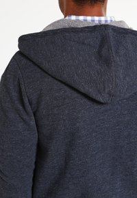 Uomo che indossa un felpa con cappuccio grigio scuro sopra una camicia a quadri blu e bianca, visto da dietro all'altezza della spalla.