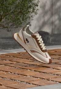 Sneaker met een bovenwerk van crème mesh en leren accenten in bruin. Heeft een witte tussenzool en een rubberen buitenzool, met een logo aan de zijkant.