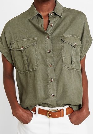 Person iført en olivengrøn kortærmet skjorte med to forlommer og hvide bukser med et brunt læderbælte.