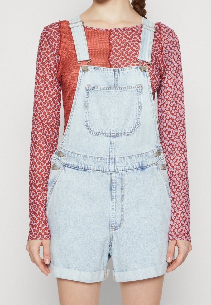 Denim-Latzshorts in Hellblau mit verstellbaren Trägern, einer Fronttasche und umgeschlagenem Saum. Darüber getragen wird ein strukturiertes, rotes, blumiges Langarmoberteil.