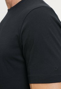 Zwarte katoenen t-shirt met korte mouwen, een ronde halslijn en subtiele stikseldetails aan de onderrand en mouwen. Bevat een klein logo op de mouw.