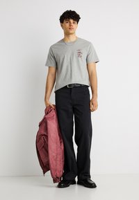 Νέος άνδρας που στέκεται, φοράει γκρι μπλουζάκι με τύπωμα, μαύρο παντελόνι, μαύρα παπούτσια, κρατώντας ένα μπορντό μπουφάν στο αριστερό του χέρι.