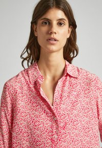 Camicia button-up rosa a fiori con colletto, motivi floreali bianchi e vestibilità rilassata. Tessuto morbido con una texture leggera.