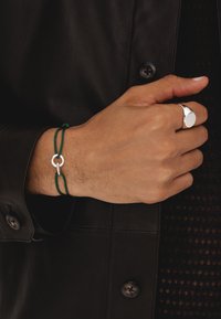 Anello circolare in argento su una mano con bracciale in corda verde che presenta due anelli metallici interconnessi, contro uno sfondo scuro e strutturato.