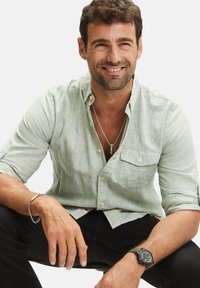 Lichtgroen, korte mouwen, knoopshirt gemaakt van een gestructureerde stof, open gedragen over een zilveren ketting, gecombineerd met zwarte broeken en een horloge.