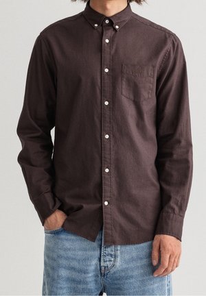 Mężczyzna w ciemnobrązowej koszuli na długi rękaw z kieszenią na piersi i jasnoniebieskich dżinsach, ręce luźno opuszczone, na jednolitym tle.