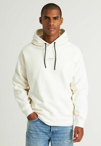 Crème hoodie met een capuchon met trekkoord, voorzien van een klein bedrukt logo op de borst. Gekleed met lichtblauwe versleten jeans.
