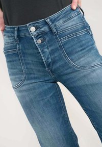 Jean en denim slim fit dans un lavage bleu moyen avec deux poches avant, une taille haute et une fermeture à double bouton. Texture lisse avec un minimum de décoloration.