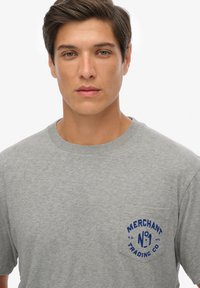 Camiseta gris de manga corta hecha de algodón suave, que presenta un logo de bolsillo azul con el texto "MERCHANT TRADING CO" y "N°1."