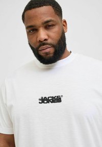 Bijela pamučna majica s kratkim rukavima, koja na prsima ima crni stilizirani logo "JACK & JONES". Klasičan okrugli dekolte.